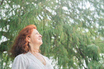 Portrait of a latin senior red-haired woman smiling looking up daydreaming in nature. Bright composition with copy space. Concept: peace of mind and happiness enjoying leisure time during retirement.