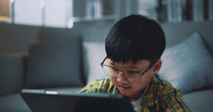 Handheld shot, Asian boy wearing glasses schoolboy learning online with a digital tablet and writing on worksheets at home. Education, e-learning and lifestyle concepts.