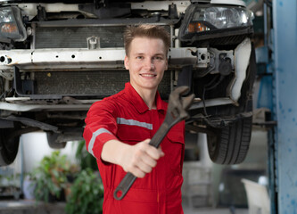 Auto mechanic in red uniform standing at car repair station holding wrench prepare to fix broken engine vehicles at mechanics garage. Smiling maintenance engineer happy work in automotive industry