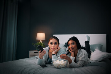Girls night, young women and popcorn while watching television on a bed in bedroom. Relaxing or...