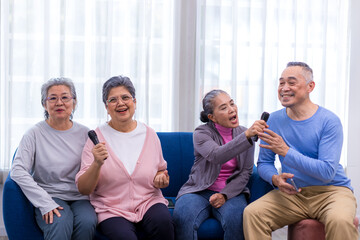 Fototapeta premium Happy senior Asian friends singing karaoke on the sofa in the living room with happy smiling face. Elderly people singing karaoke. Friends singing karaoke at home