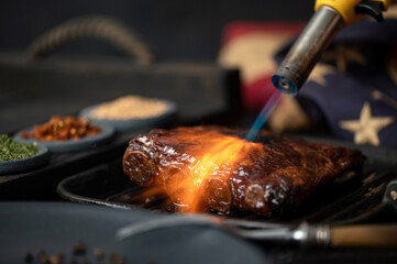 Grilled ribs under stream of fire. Grilled ribs in pan. Plates of spices and American flag, fork on table. American cuisine. Dark wooden background. Side view. 