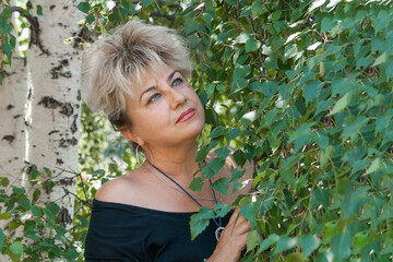 55-60-year-old woman stands against the background of birch branches. Large portrait of a Russian woman with blonde hair