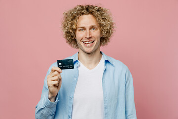 Young smiling happy fun caucasian blond man wears blue shirt white t-shirt hold in hand mock up of credit bank card isolated on plain pastel light pink background studio portrait. Lifestyle concept.