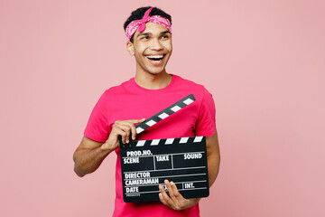 Young man of African American ethnicity wear headscarf t-shirt casual clothes hold in hand classic black film making clapperboard isolated on pastel plain light pink color background studio portrait.