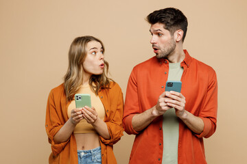 Young shocked amazed fun couple two friend family man woman wear casual clothes hold use mobile cell phone together look to each other isolated on pastel plain beige color background studio portrait