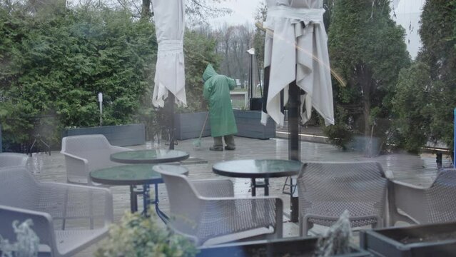 male figure man in green rain coat sweeping cleaning cafe terrace broom rainy weather day. working man in green hood cleanup dirty floor outdoors stormy weather cloudy daytime. person occupation