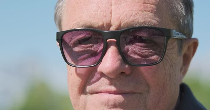 Elderly man in sunglasses looks at the camera and smile. Close-up face, portrait, front view. Sunny day, outdoors
