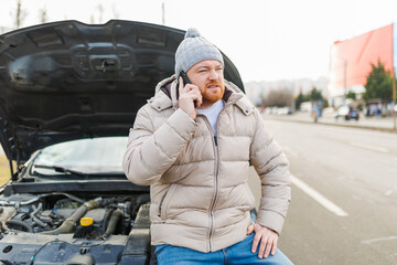 A young man looks angry and frustrated near his broken car and speaks on his mobile phone,