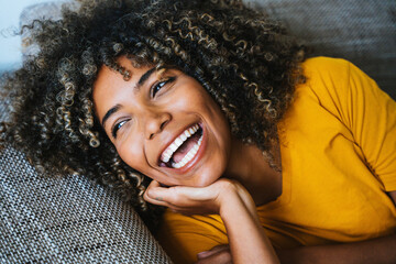 Happy black woman relaxing lying on the sofa at home - Portrait of comfortable girl laughing on the couch - Healthy lifestyle concept with young female smiling at camera