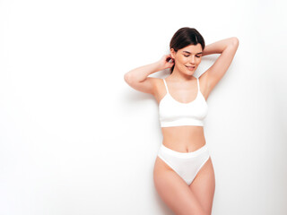 Fototapeta premium Portrait of young beautiful sexy woman in white lingerie. Smiling carefree model wearing pure underwear. Hot brunette posing near wall in studio interior. Perfect body. Slim and fit