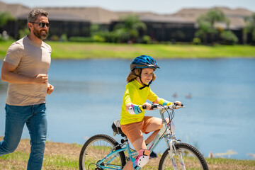 Obraz premium Happy Fathers day. Father and son in bike helmet for learning to ride bicycle at park. Father helping son cycling. Father and son on the bicycle on summer day. Kid son trying to ride bike with father.
