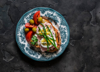 Sandwich with boiled egg, ham, asparagus and tomatoes, olives in a plate on a dark background, top view