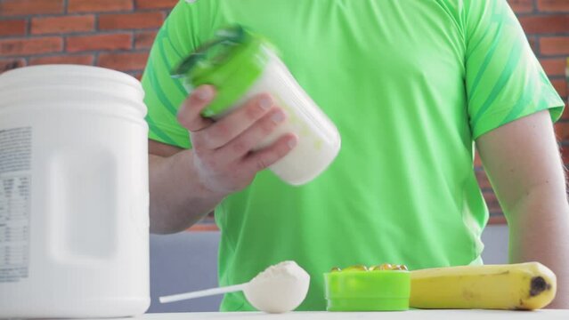 Unrecognizable man in a green sport shirt shaking protein power drink, process of making protein milk banana smoothie, healthy sport nutrition, crop image
