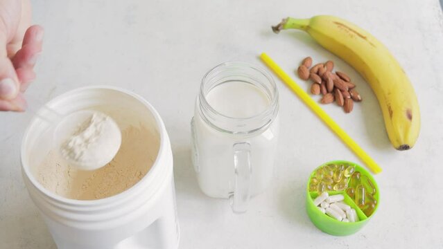 Male hand puts scoop with white whey or soy protein powder from a jar into glass with a milk, process of making banana smoothie drink, sport nutrition