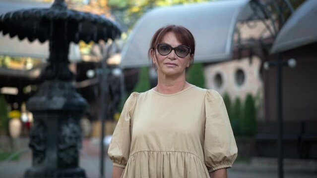 Zoom out portrait of confident beautiful lady in eyeglasses and dress standing at fountain in city park looking at camera. Gorgeous Caucasian woman posing outdoors