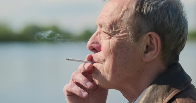 Elderly man smokes thin cigarette outside in park. Releases smoke through mouth. Side view, close-up, portrait.