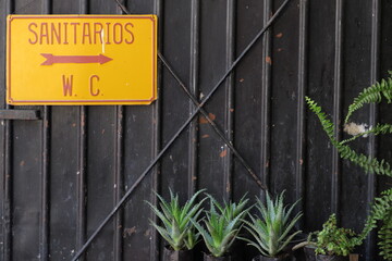 Sanitarios WC (sanitaire WC). Panneau jaune. Lettrage rouge. Sur cloison de métal noire. Plantes...