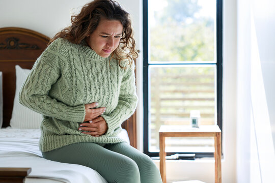 Young Woman Sitting On The Bed At Home With A Pain In Her Stomach