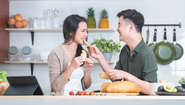Attractive Asian young sweet couple cooking together in home kitchen. Handsome man smiling feeding his beautiful woman with bread while preparing meal, fresh fruits for breakfast. Family joyful moment