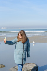 Smiling young woman in blue parka walking near sea at sunny spring day. Taking selfie