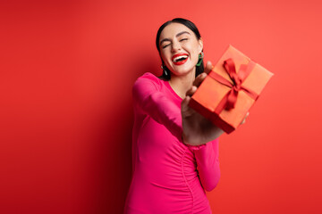 excited woman with brunette hair and trendy earrings smiling while standing in magenta party dress and holding wrapped gift box for holiday on red background