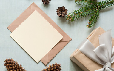 Christmas still festive life. Blank greeting card, invitation mockup, envelope and gift box with ribbon on textile background. Fir branches and cones on the table. Copy space, top view.