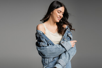 alluring young woman with brunette hair and natural makeup standing in blue jeans and fashionable denim jacket while smiling and posing isolated on grey background