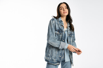 fashionable young model with brunette hair and flawless makeup posing in blue denim jacket and jeans while standing and looking at camera on white background