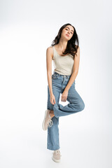 full length of alluring woman with shiny brunette hair, natural makeup and perfect skin posing in tank top and touching denim jeans on white background