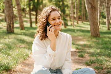 Cheerful curly woman is having a phone call while enjoying sitting in a forest after work.