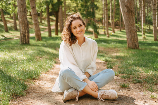 Smiling curly woman wearing white shirt is sitting in lotus pose in forest on a sunny day.