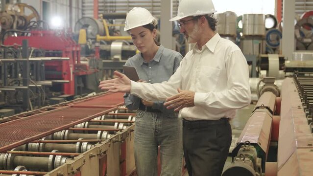  male and female engineer, both wearing safety helmets, discuss the operations of machinery in a large industrial factory.
