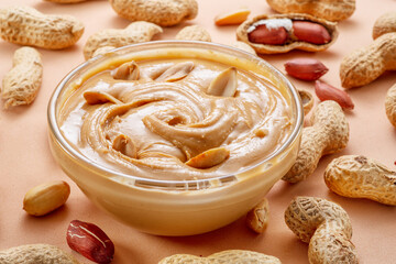 Bowl of peanut butter and peanuts around it on beige background.