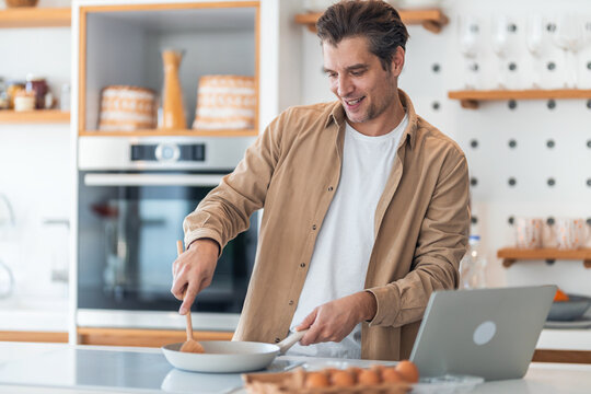 A smiling male freelancer making scrambled eggs in a pan and working on a laptop.