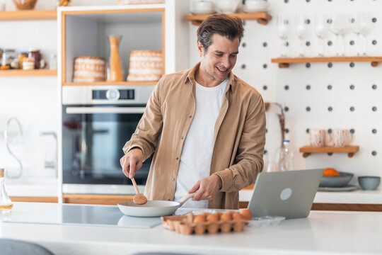 A smiling male freelancer making scrambled eggs in the pan during work time on the laptop.