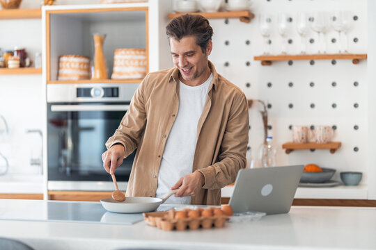 A smiling male freelancer making himself fried eggs for breakfast and working over the laptop.