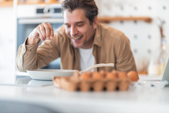 A smiling male freelancer salts eggs in the pan for breakfast during the work break.