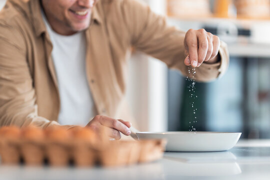 A smiling man makes scrambled eggs and salts them while they are in a pan.