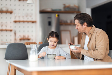 A  father holding a bowl of cereal with milk and looking at a hungry daughter.