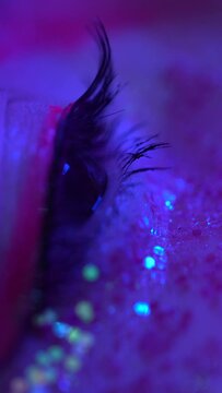 Extreme closeup view of female eye with shiny make-up with sparkles and arrows. Cinematic macro shot of female eye illuminated pink-blue neon light. Selective focus, handheld. Vertical video shot