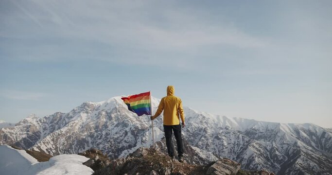 Caucasian Young Man Standing on the Peak, Holding the Rainbow LGBTQ Flag on Top of Mountains. Male GenZ Activist Celebrating Love, Freedom, Equality and Diversity, Enjoying an Beautiful Sunset View