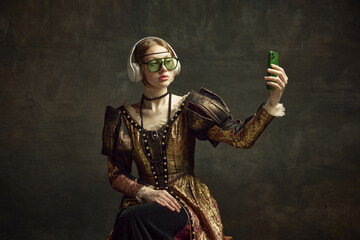 Portrait of young beautiful girl in vintage dress, trendy sunglasses and headphones, taking selfie with phone against dark green background. Concept of history, renaissance art, comparison of eras - obrazy, fototapety, plakaty