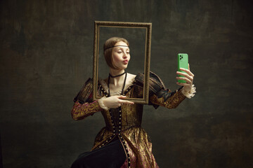 Portrait of pretty young girl, royal person in vintage dress holding picture frame and taking selfie with phone against dark green background. Concept of history, renaissance art, comparison of eras - obrazy, fototapety, plakaty