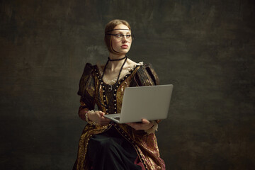 Portrait of young pretty girl, queen in vintage dress posing with laptop over dark green background. Education, freelance job, business. Concept of history, renaissance art remake, comparison of eras - obrazy, fototapety, plakaty