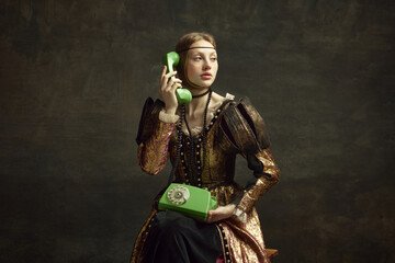 Portrait of young girl in vintage dress, royal person talking on retro phone against dark green background. Communication. Concept of history, renaissance art remake, comparison of eras