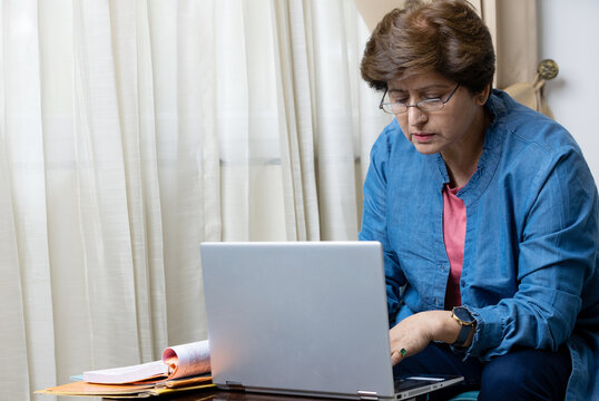 Focused and confident senior woman in glasses using a laptop for remote work sitting at the desk at home. Concentrated aged woman typing mails answers, messaging online