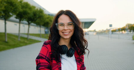 Obraz premium Front view of attractive positive emotional cheerful 25-aged brunette with long wavy hair in glasses and with headphones around her neck which poses on the background of city airport