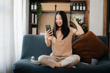 Obraz premium Attractive happy young Asian student studying at the college library, sitting at the sofa, using a laptop computer, tablet and headphones having a video chat.