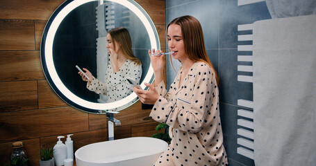 Young happy smiling woman is using smartphone for work or entertainment while brushing her teeth with toothpaste in bathroom at home.